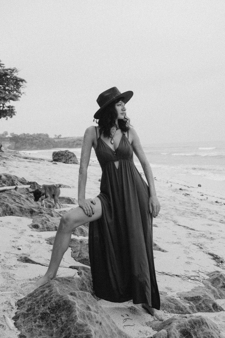 Black and white portrait of a woman in a wide-brimmed hat standing barefoot on a rocky beach, one foot grounded on stone
