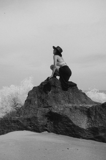 Black and white photo of a woman crouched on a rocky outcrop above crashing waves