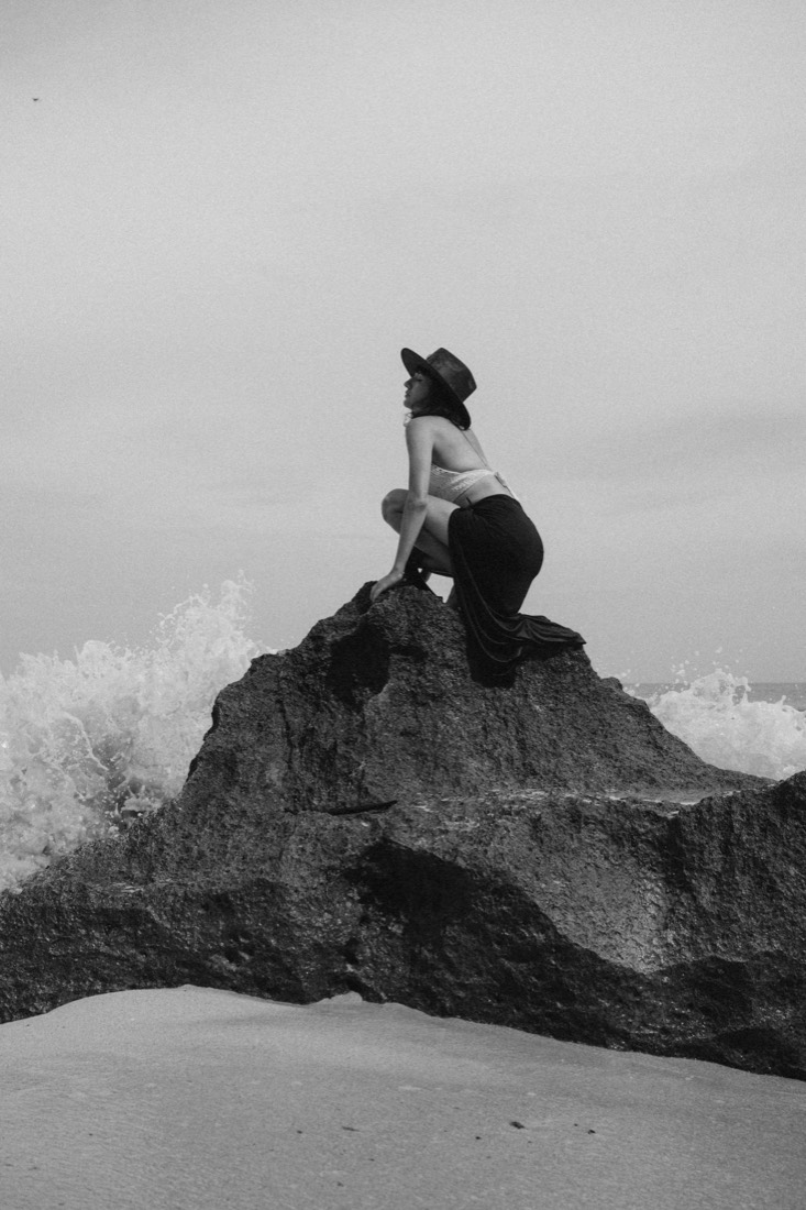 Black and white photo of a woman crouched on a rocky outcrop above crashing waves
