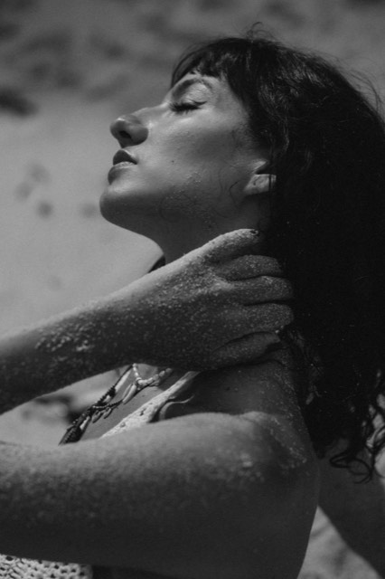 Black and white close-up photo of a woman with sand on her skin, hands at her collarbone, eyes closed